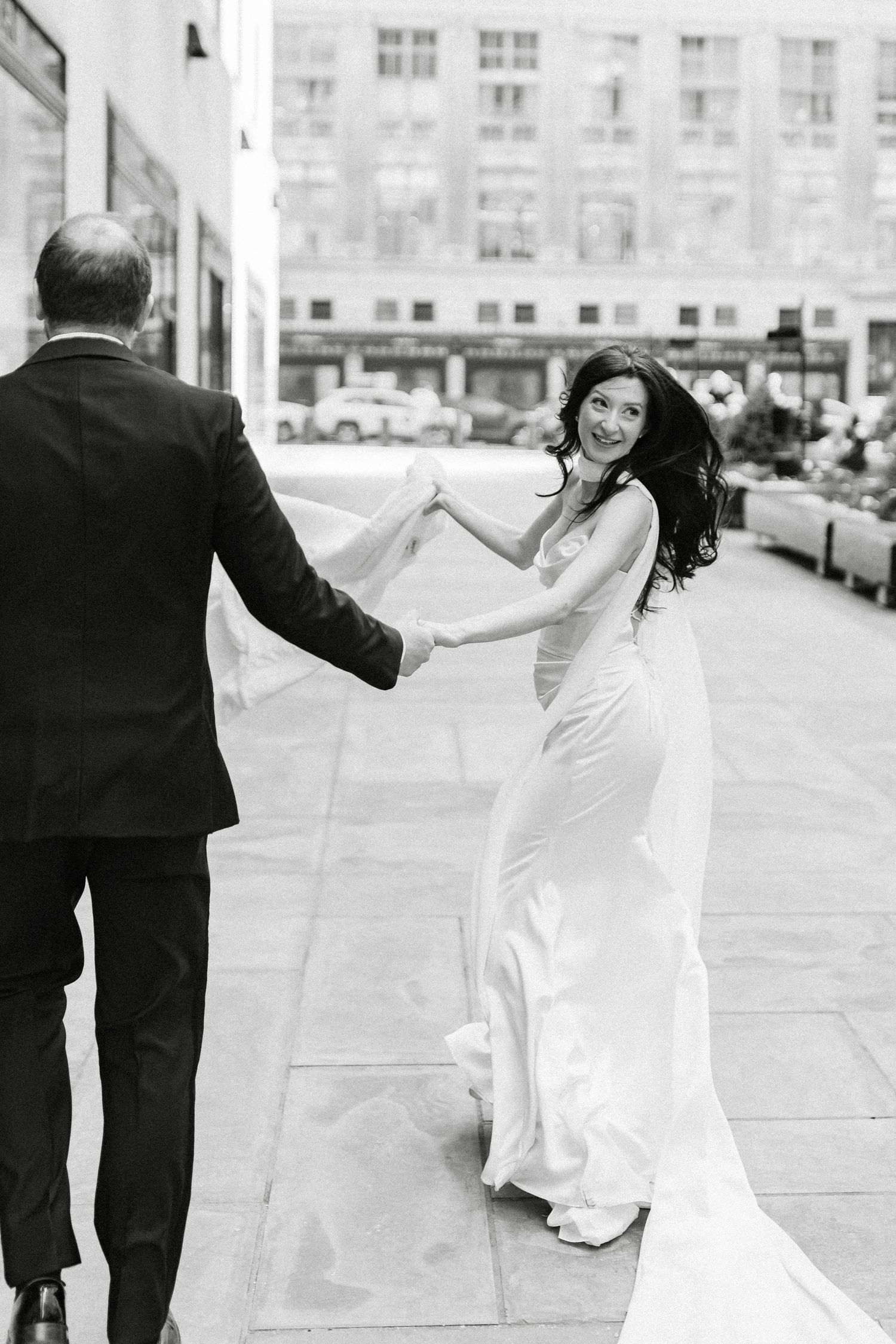 Black and white photo of a smiling bride in a sleek gown holding hands with a groom while walking on a city sidewalk.