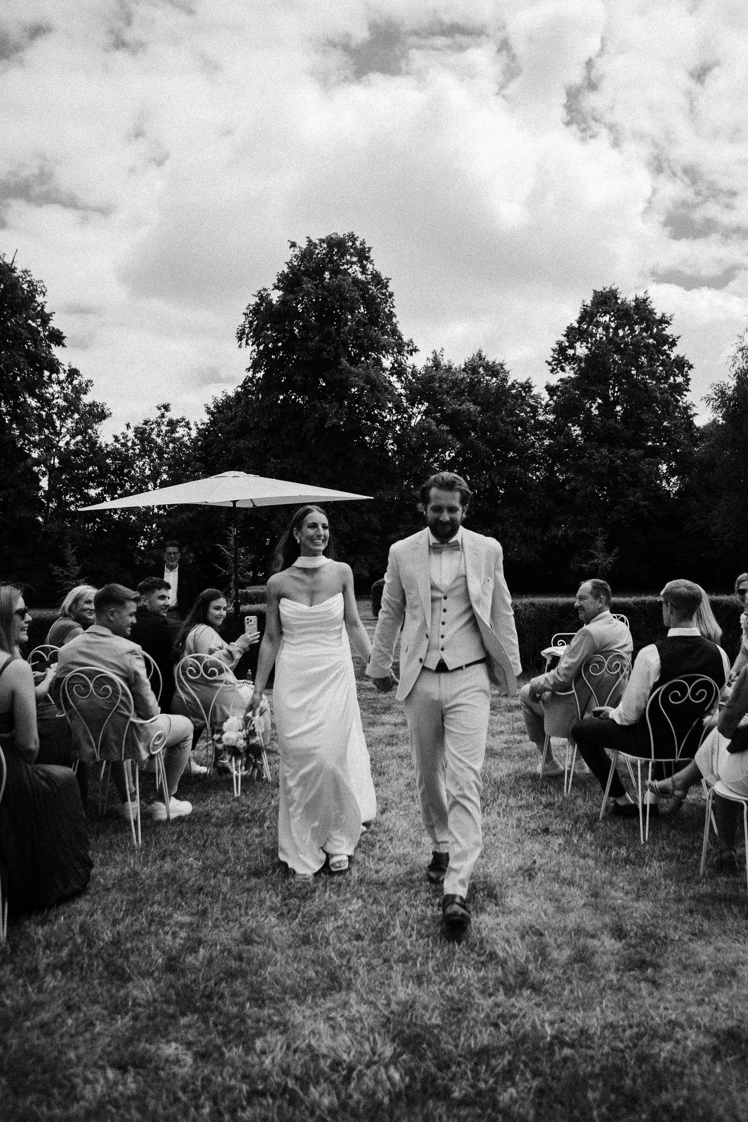 Black and white photo of a smiling bride and groom walking hand in hand down the aisle at an outdoor wedding ceremony.