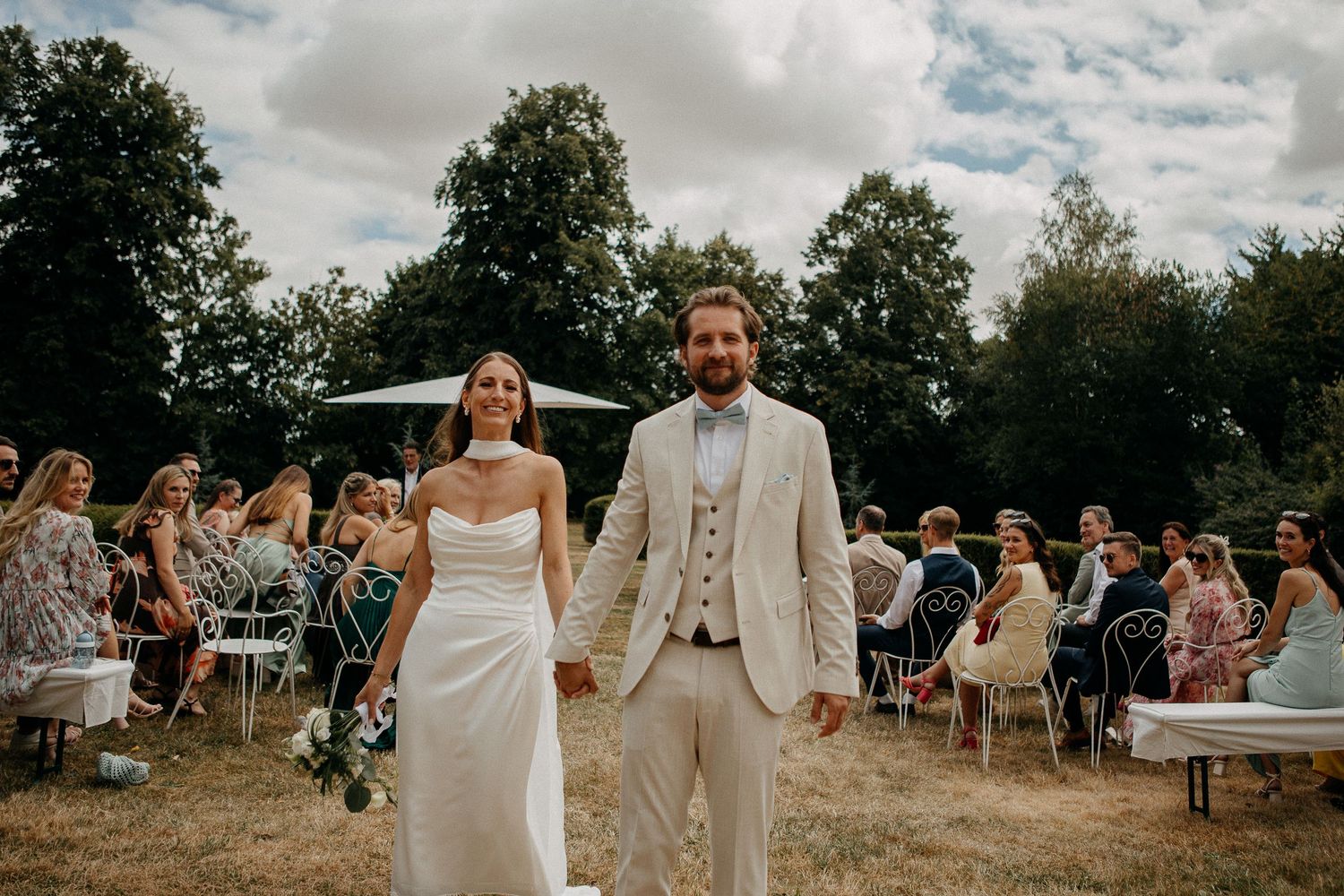 Bride and groom holding hands walking down the aisle at an outdoor wedding ceremony with guests seated on chairs.