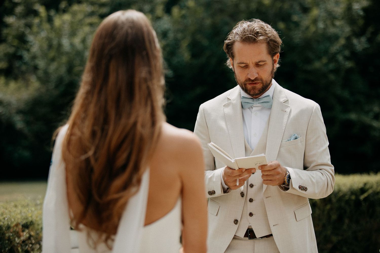 Groom in beige suit and blue bow tie reads wedding vows from paper during outdoor ceremony.