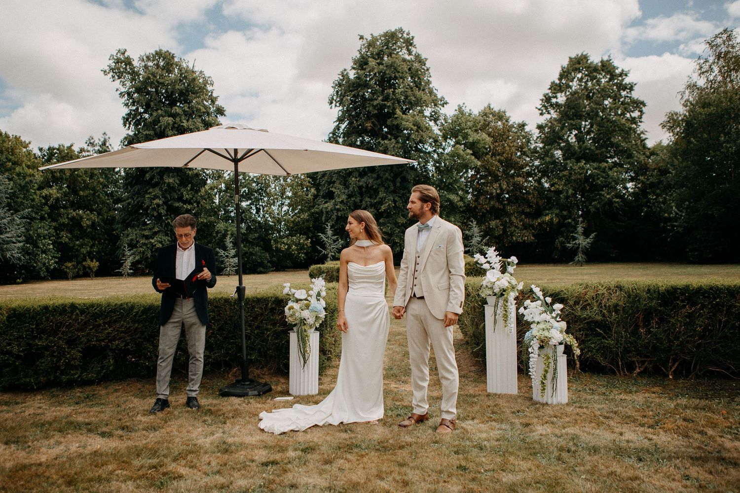 Outdoor wedding ceremony with bride and groom holding hands, officiant, white floral pillars, and large umbrella on lush green lawn.