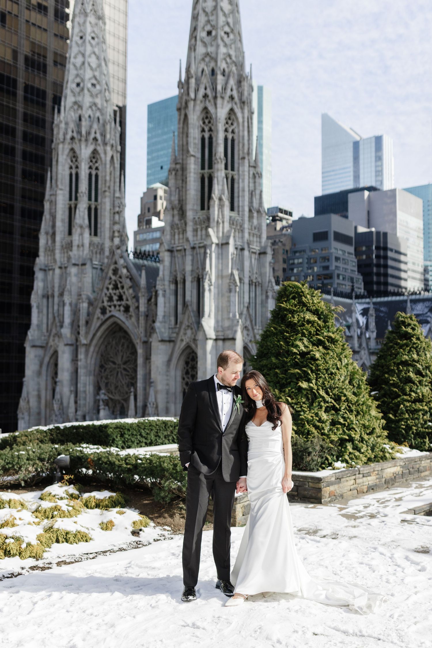 Bride and groom pose in winter snow before St. Patrick's Cathedral spires and NYC skyline on their wedding day.
