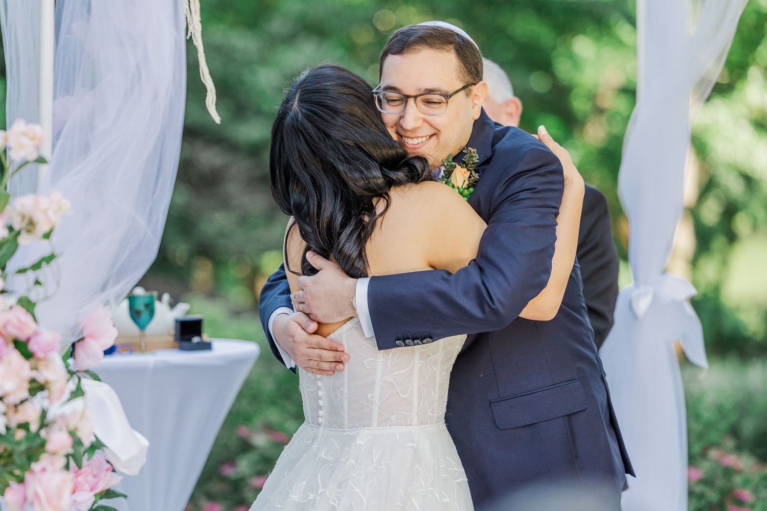 Bride and groom share a heartfelt hug during their outdoor wedding ceremony surrounded by pink flowers.
