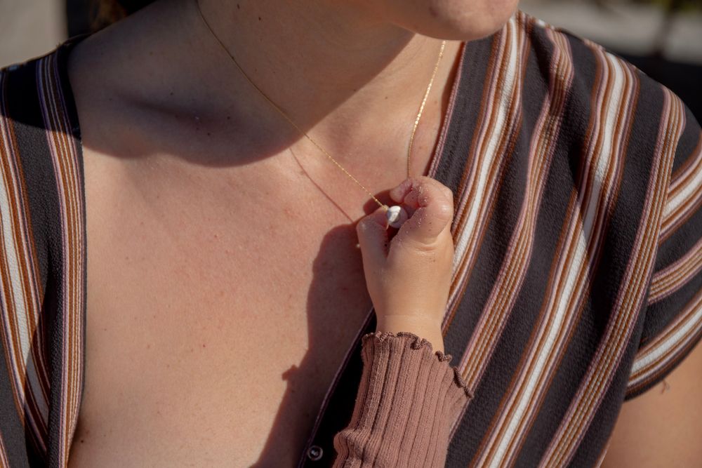 Woman in striped top holding a delicate gold chain necklace pendant close to her chest in sunlight.