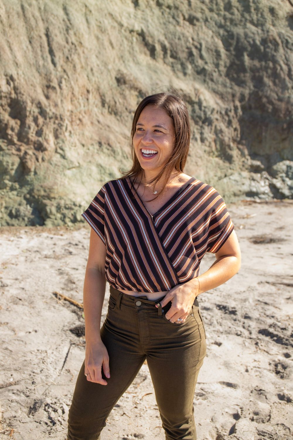 A laughing woman in a striped top and olive pants stands on a sandy beach with rocky cliffs behind her.