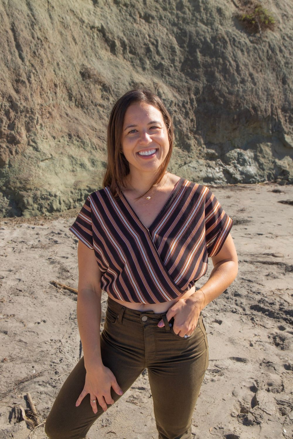 Smiling woman in striped crop top and olive jeans poses on sandy beach with rocky cliffs in background.