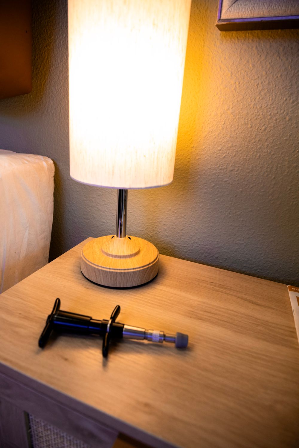 A syringe on a wooden nightstand beside a glowing bedside lamp in a dimly lit bedroom.