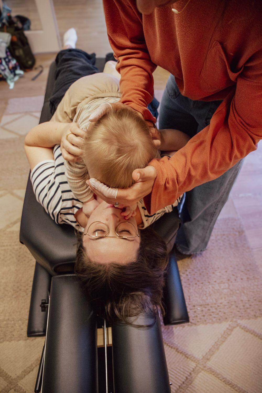 Chiropractor performing a gentle neck adjustment on a patient lying on a treatment table with a baby nearby.