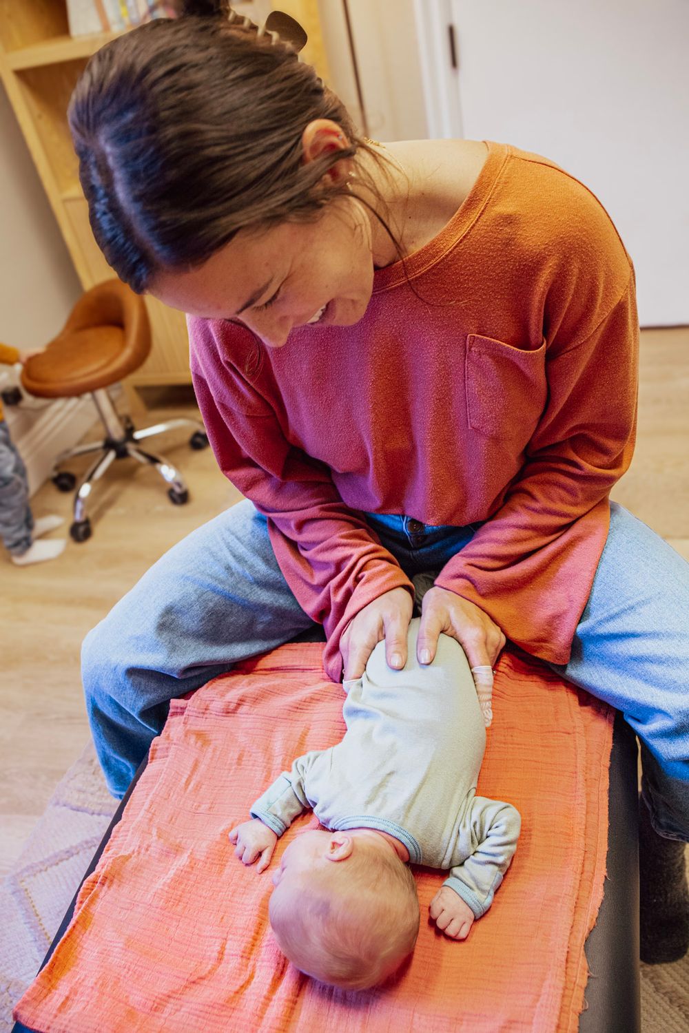 A woman in an orange top gently massages a baby lying on a pink blanket on a table indoors.