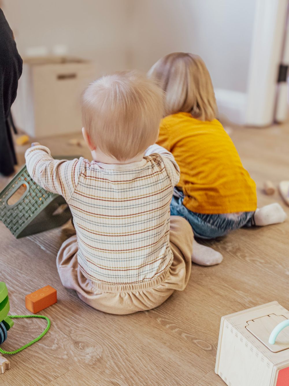Two young children sitting on hardwood floor playing with colorful wooden toys at home.