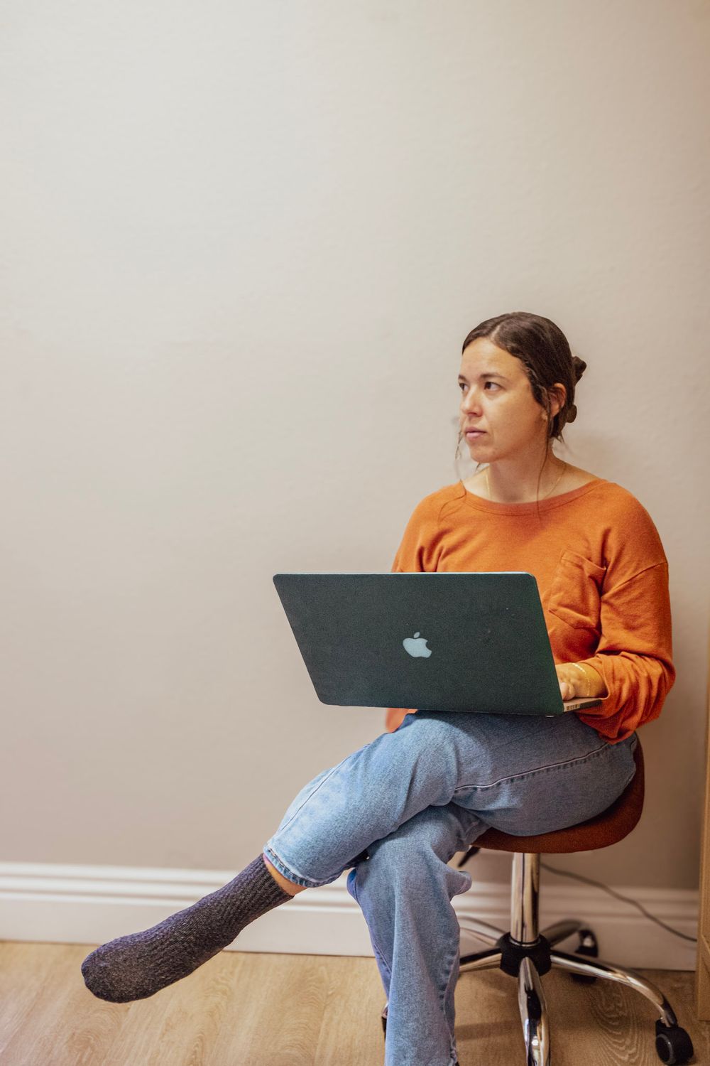 Young woman in orange sweater sitting on chair using Apple MacBook laptop computer.