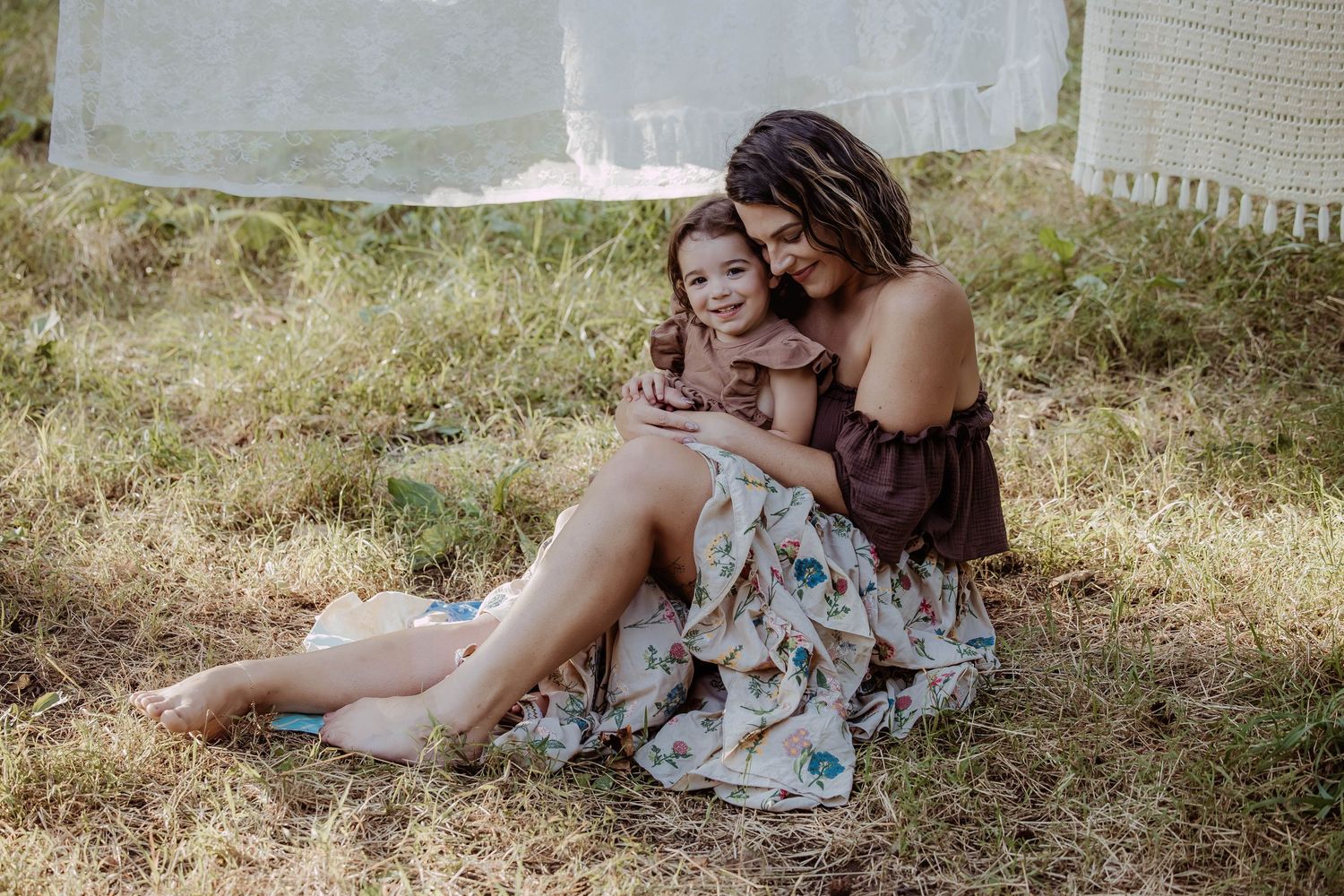 A mother and young daughter share a tender embrace sitting on grass outdoors in boho floral dresses.
