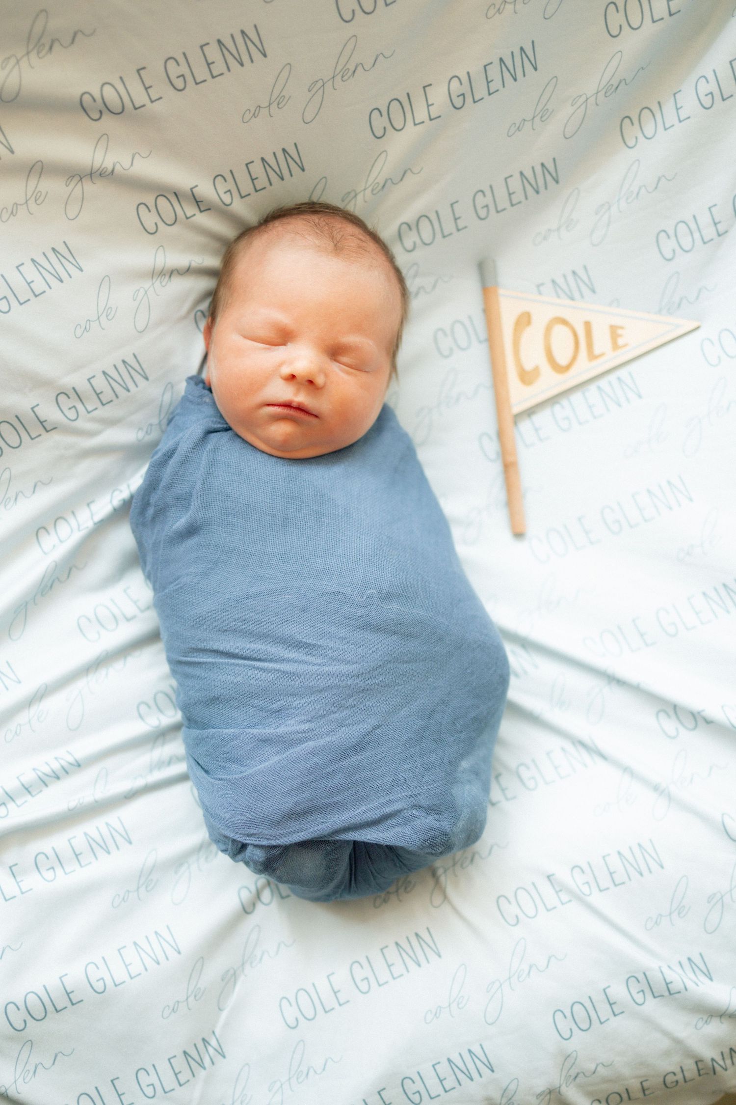 Newborn baby named Cole Glenn swaddled in blue wrap, sleeping peacefully on personalized name blanket with pennant flag.
