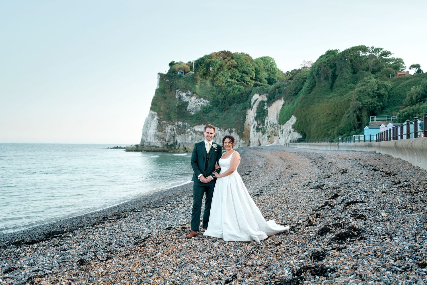 Bride and groom posing on a pebble beach with white cliffs and lush greenery in the background.