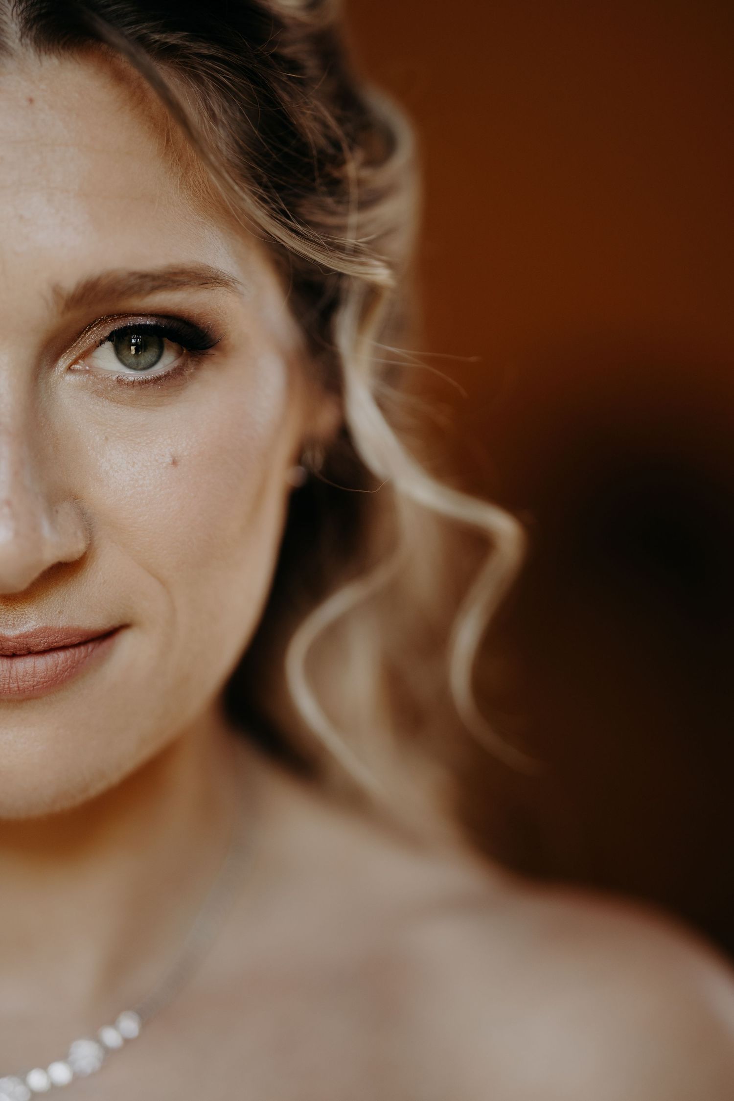 Close-up portrait of a bride with green eyes, soft curls, pearl necklace, and elegant makeup on warm brown background.