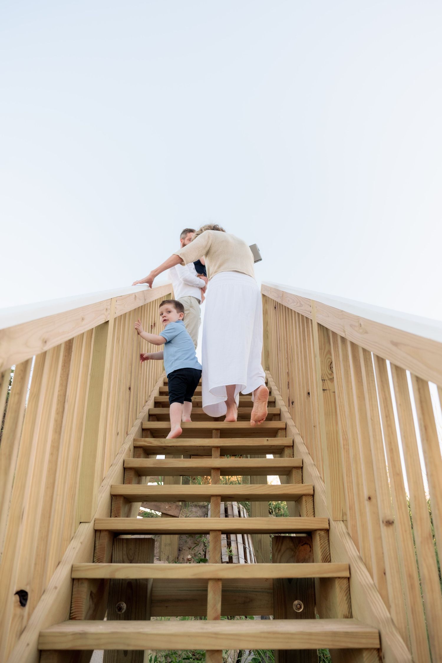 Family climbing wooden outdoor stairs together, barefoot woman in white dress leading young children up steps against clear sky.