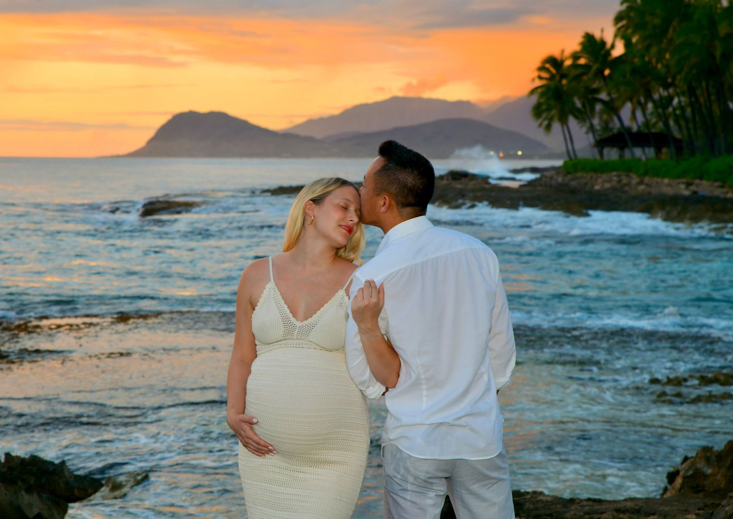 Pregnant couple sharing a romantic moment at sunset on a rocky beach with ocean waves and palm trees in Hawaii.