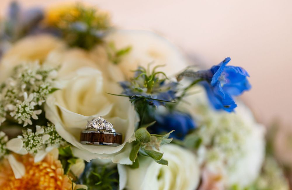 Wedding rings resting on a bridal bouquet with white roses, blue flowers, and yellow blooms.