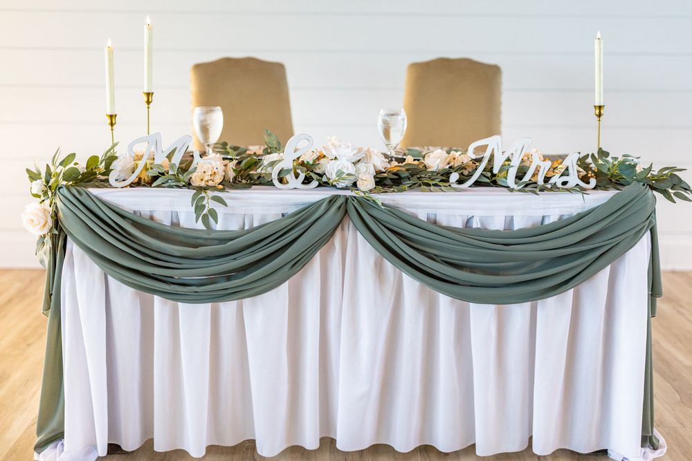Elegant wedding sweetheart table with sage green draping, floral garland, gold candles, and Mr. & Mrs. signs.