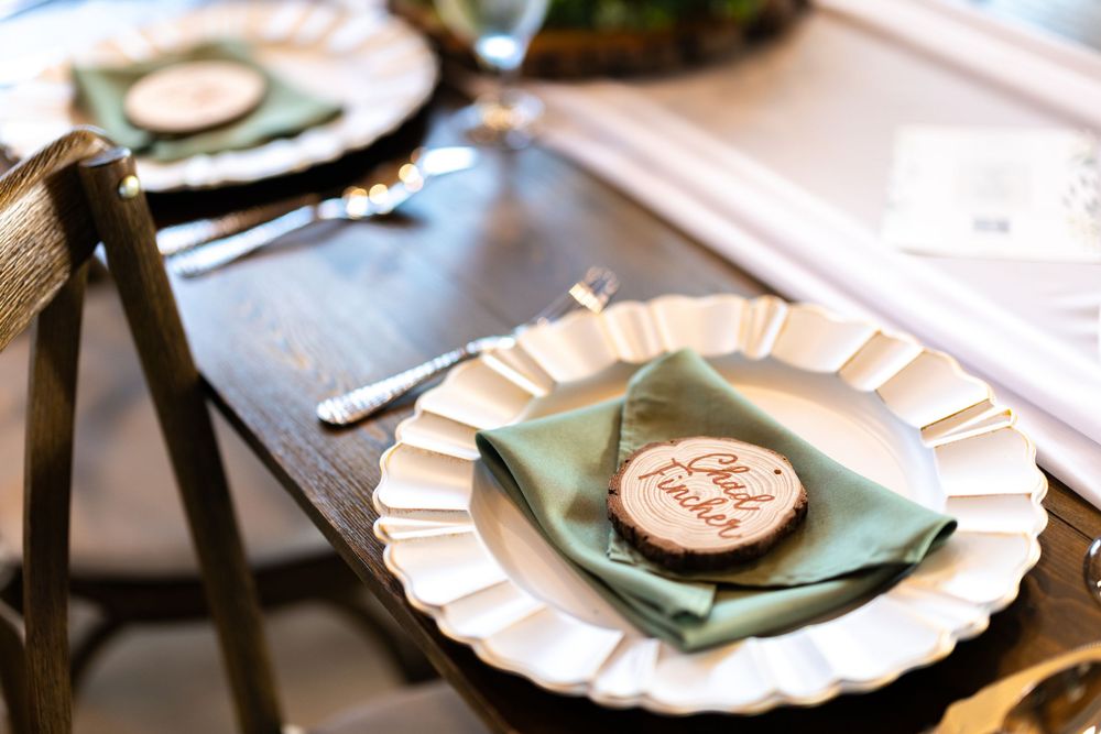 Elegant wedding table setting with white scalloped plates, sage green napkins, and rustic wood slice place cards.