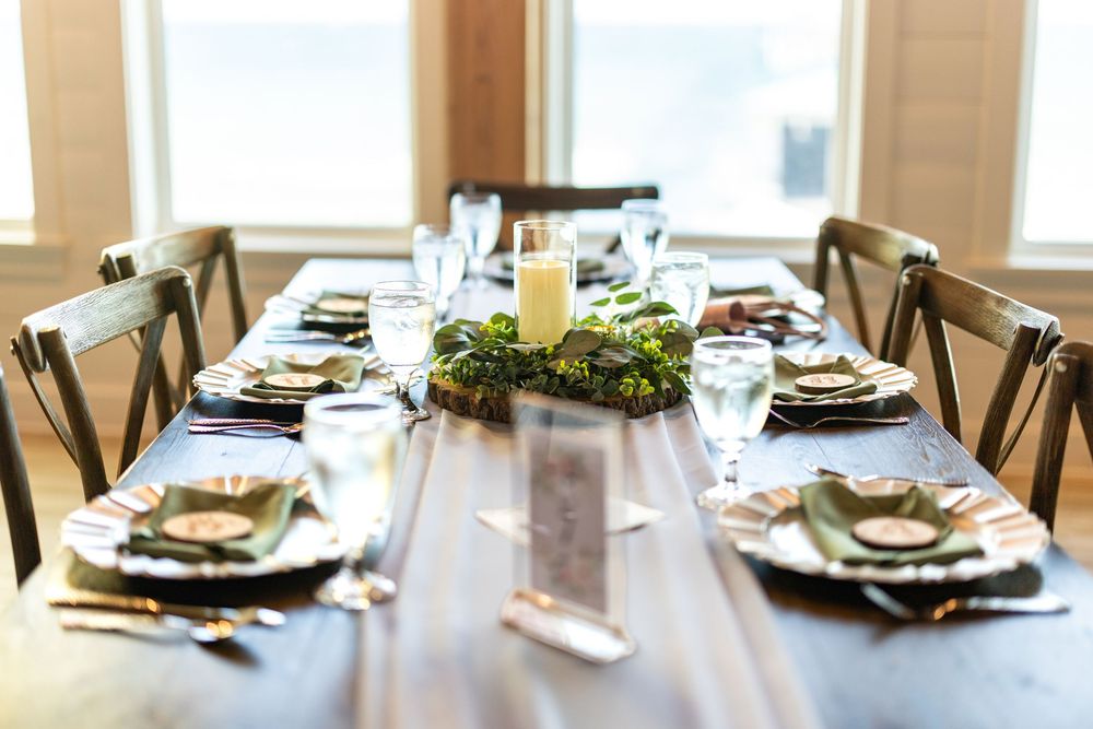 Elegant rustic dining table set for a formal dinner with candle centerpiece, greenery, and place settings by sunny windows.
