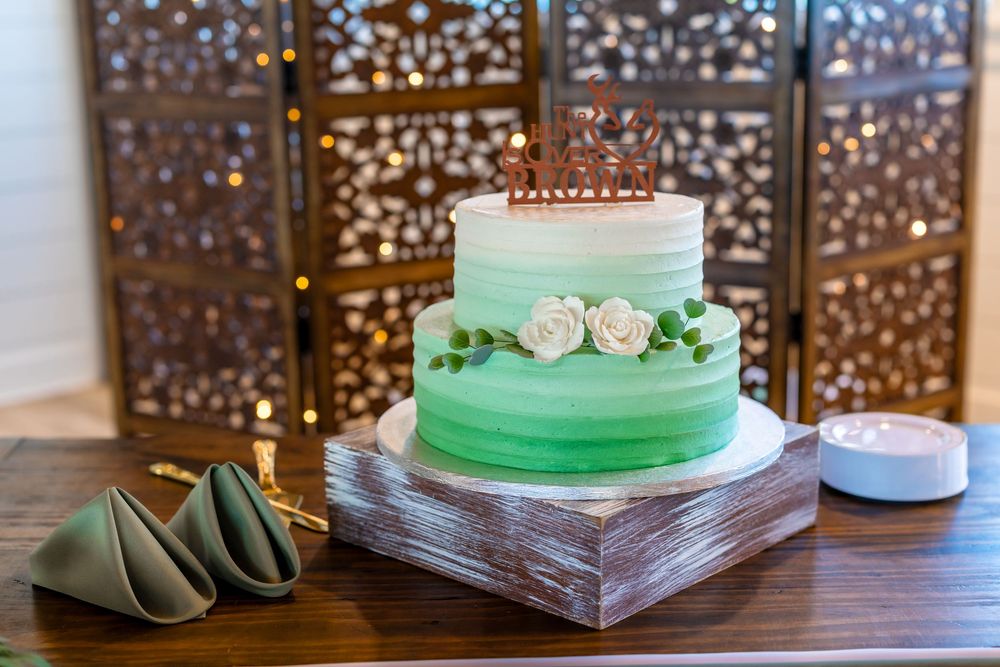 Two-tier green ombre wedding cake with white roses and couple topper on rustic wooden stand.