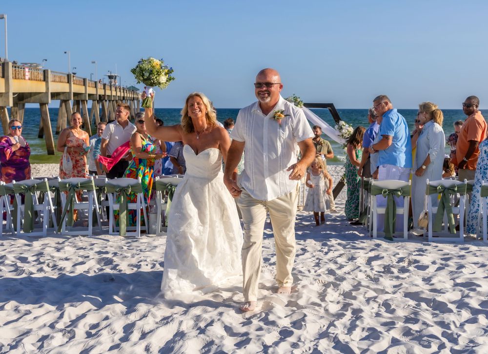 Joyful newlyweds walk down sandy beach aisle after wedding ceremony near ocean pier, bride holding bouquet.
