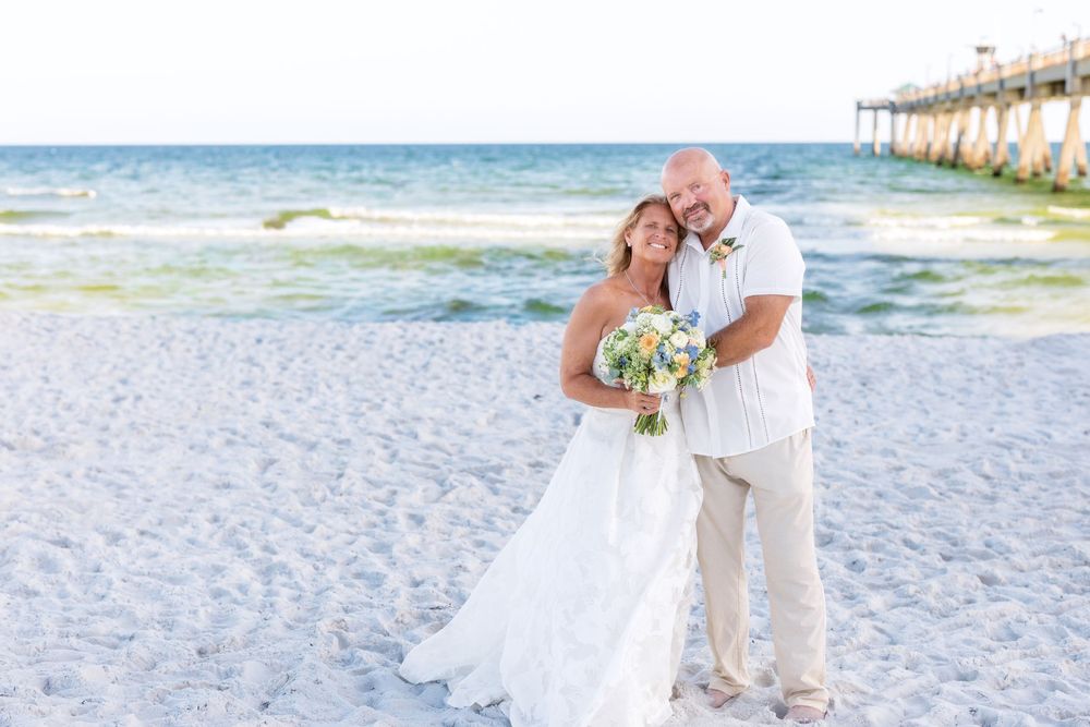 Happy married couple embraces on white sand beach near pier, bride holding blue and white bouquet.