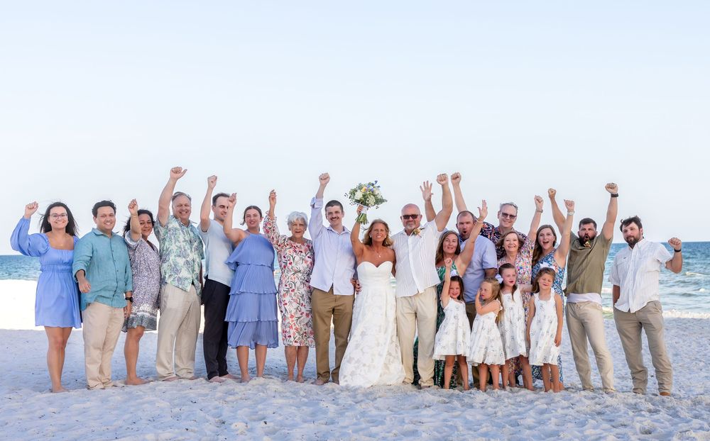 Wedding party celebrating on beach, raising hands in joy with bride and groom in center.