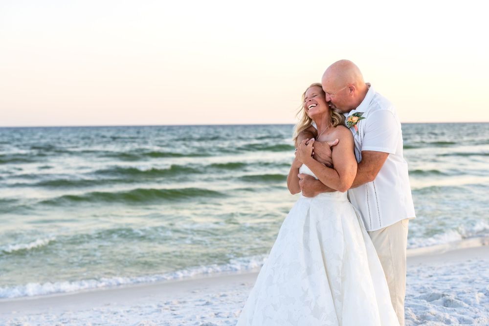 Couple embracing on a sandy beach at sunset, bride in white gown and groom in white shirt, ocean waves behind them.