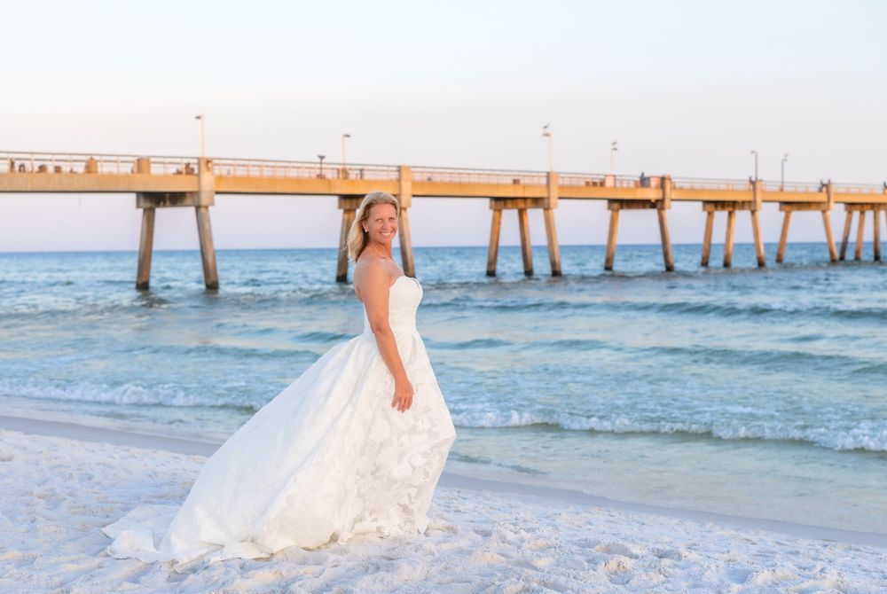 Bride in white wedding gown posing on sandy beach with ocean waves and wooden pier in background.