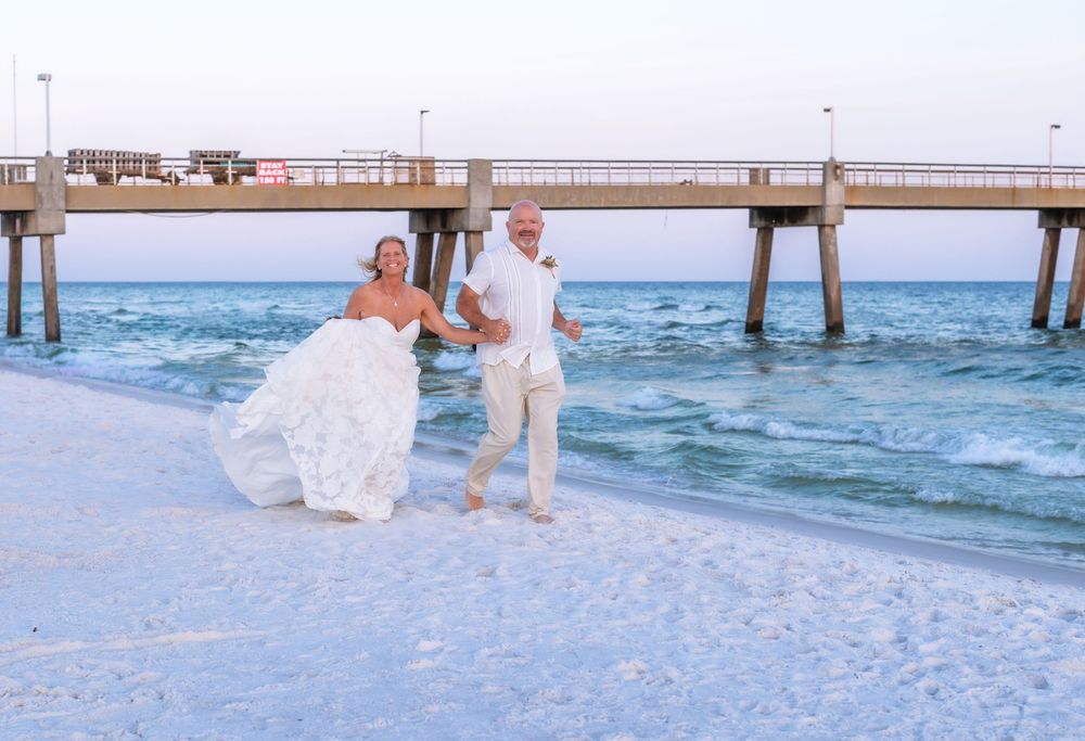 Bride and groom running joyfully on white sandy beach near pier, bride in flowing white gown, sunny day.