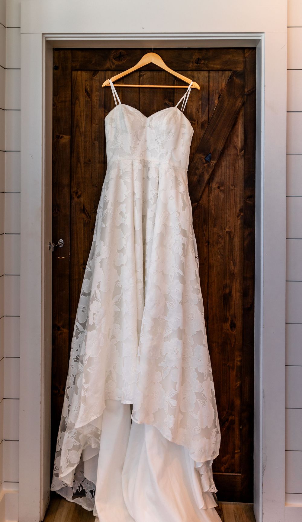 Elegant white lace A-line wedding gown with spaghetti straps hanging on wooden hanger against rustic barn door.