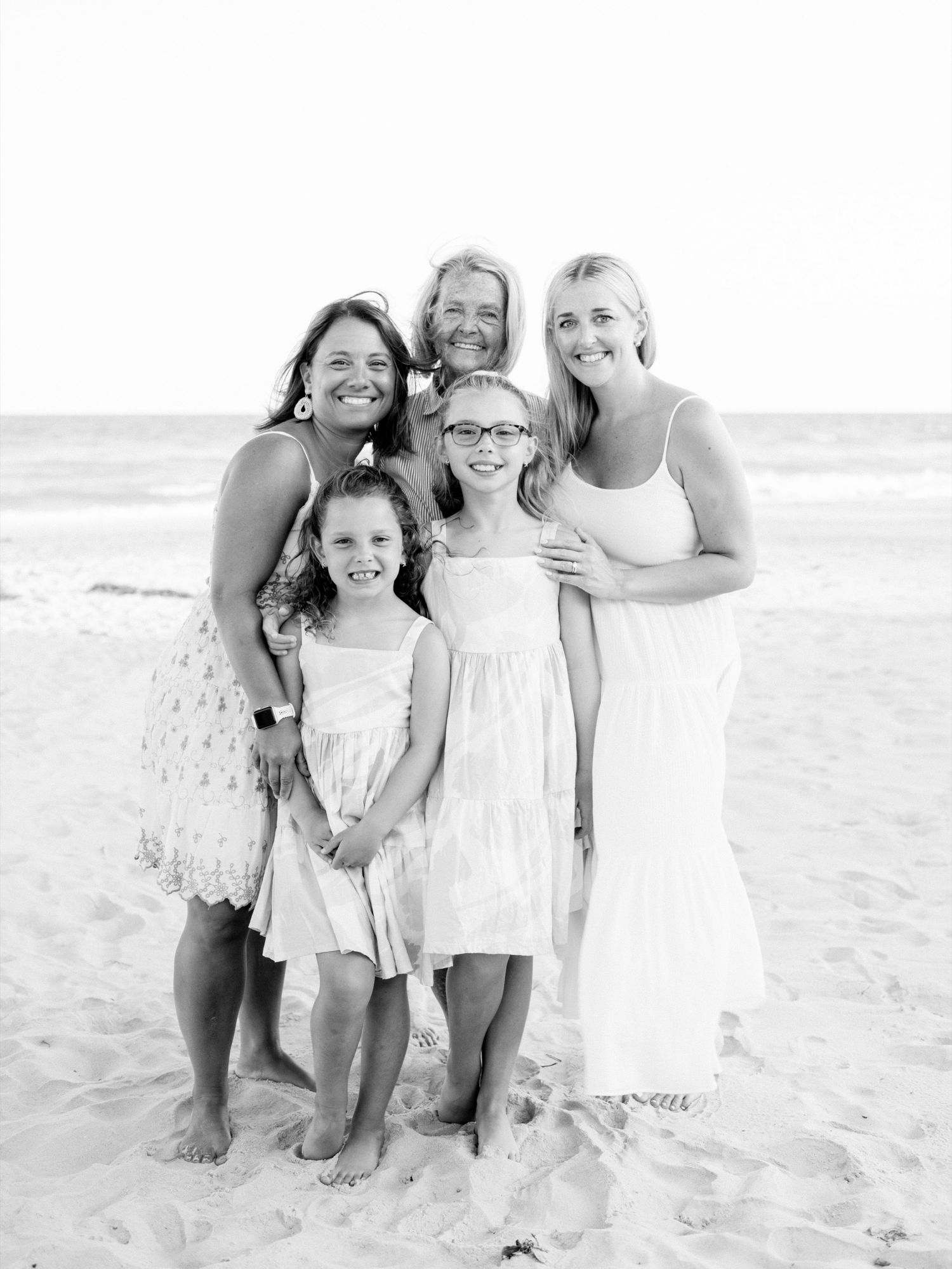 Black and white beach photo of a multigenerational family of five women and girls smiling together on sandy shore.
