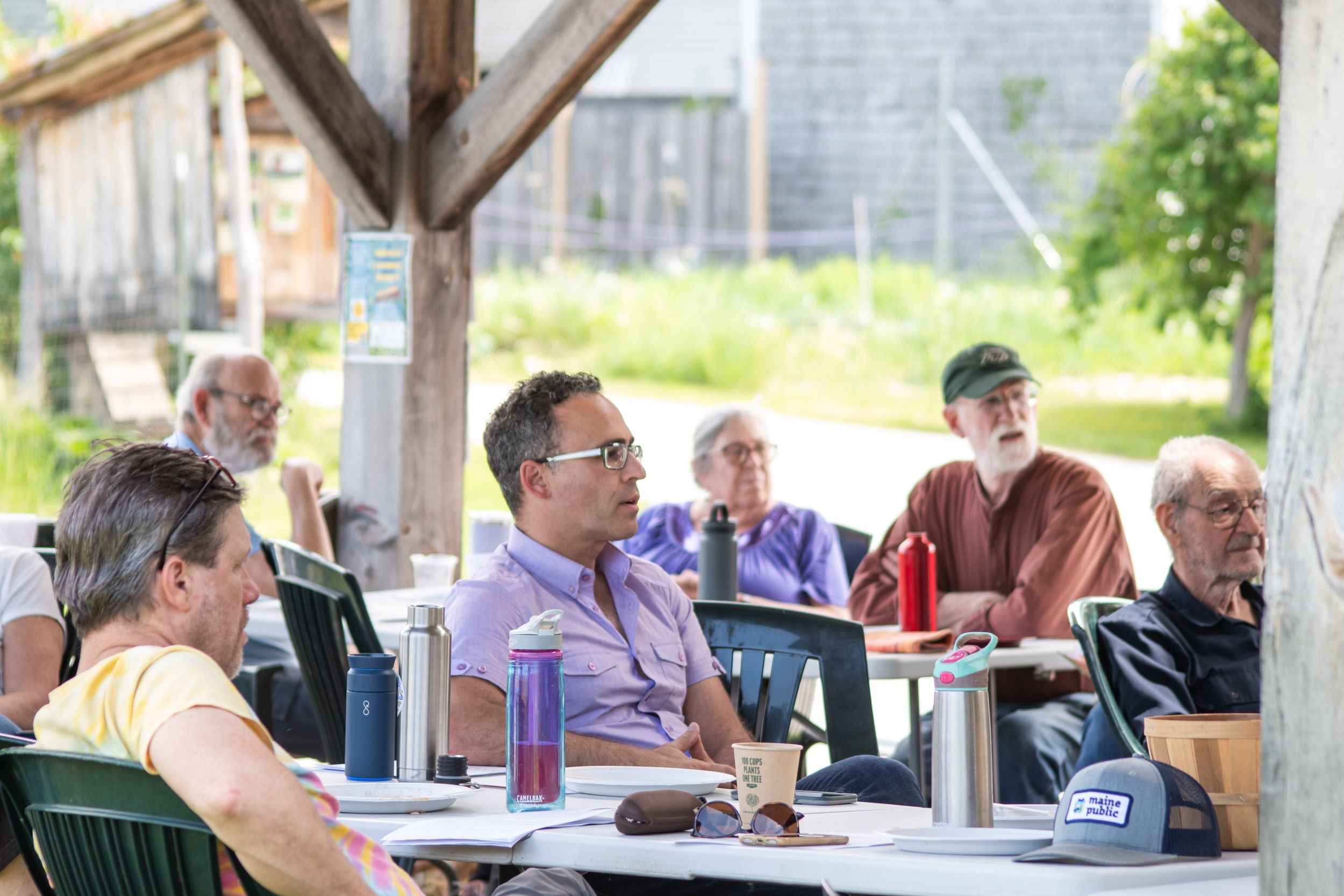Group of men seated at outdoor tables under a wooden pergola during a daytime meeting.