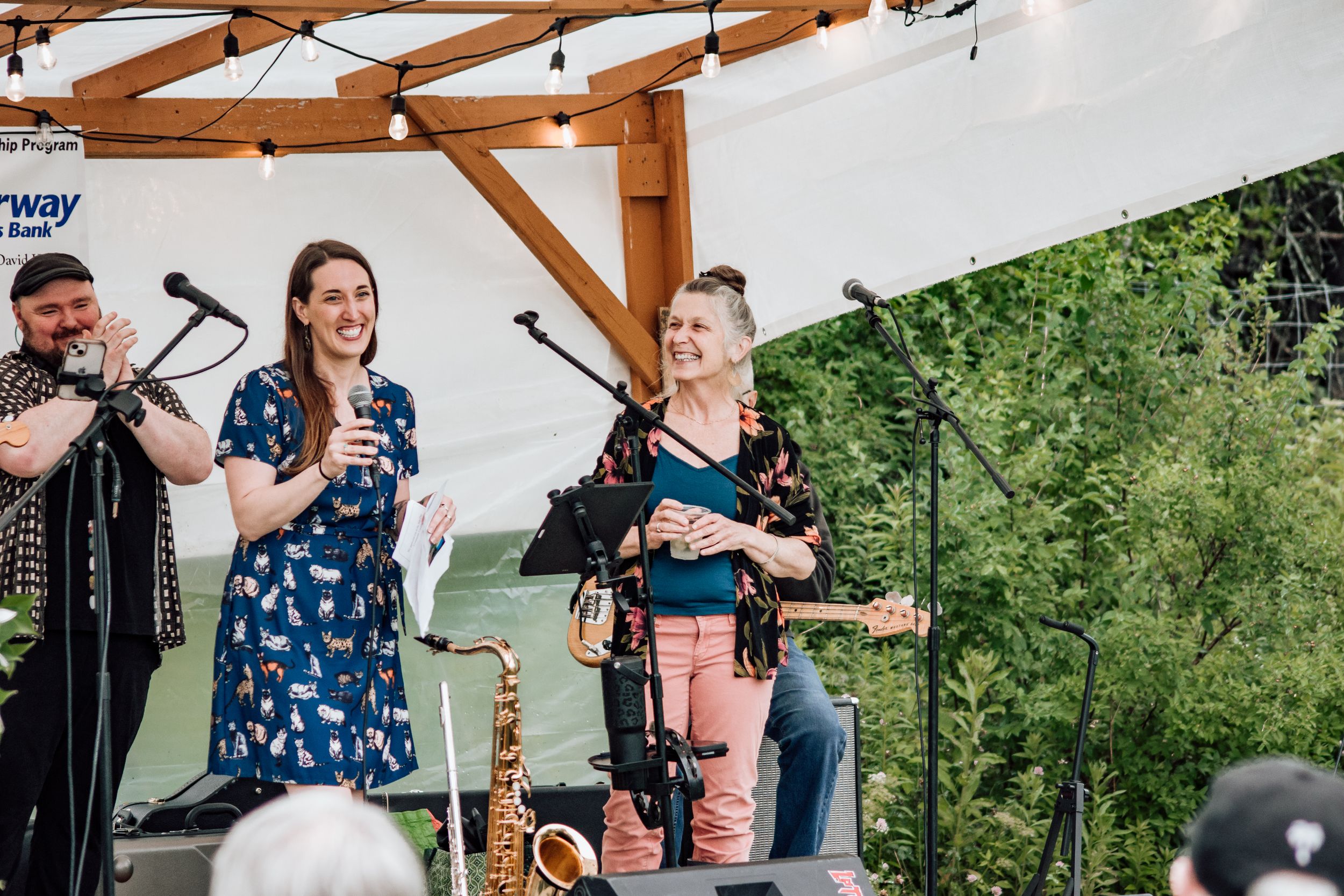 Two women performing on an outdoor stage with microphones and instruments at a summer event.