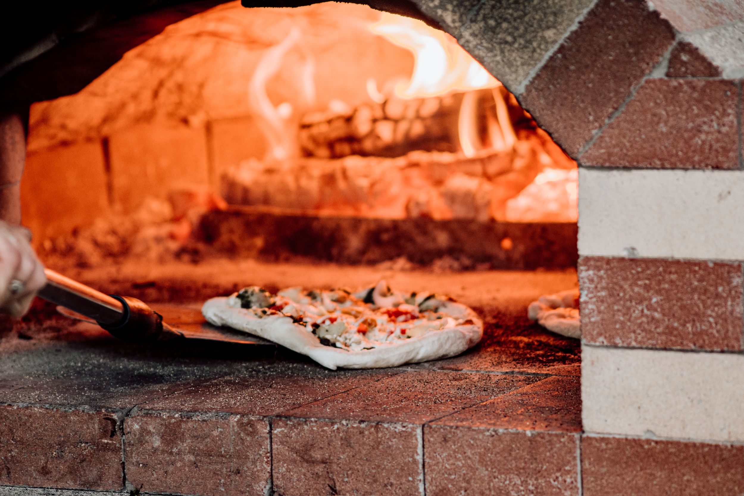 Fresh pizza being placed into a wood-fired brick oven glowing with orange flames.