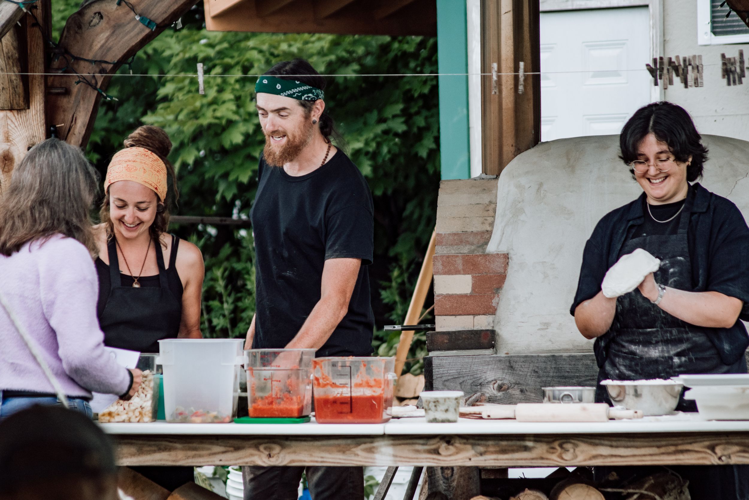 Three smiling food vendors serving guests at an outdoor pizza station with fresh ingredients.
