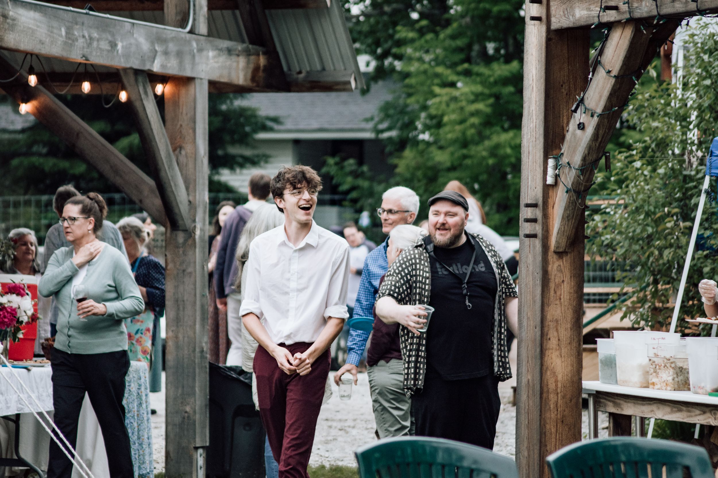 Two young men laughing and socializing under a wooden pergola at an outdoor evening gathering.