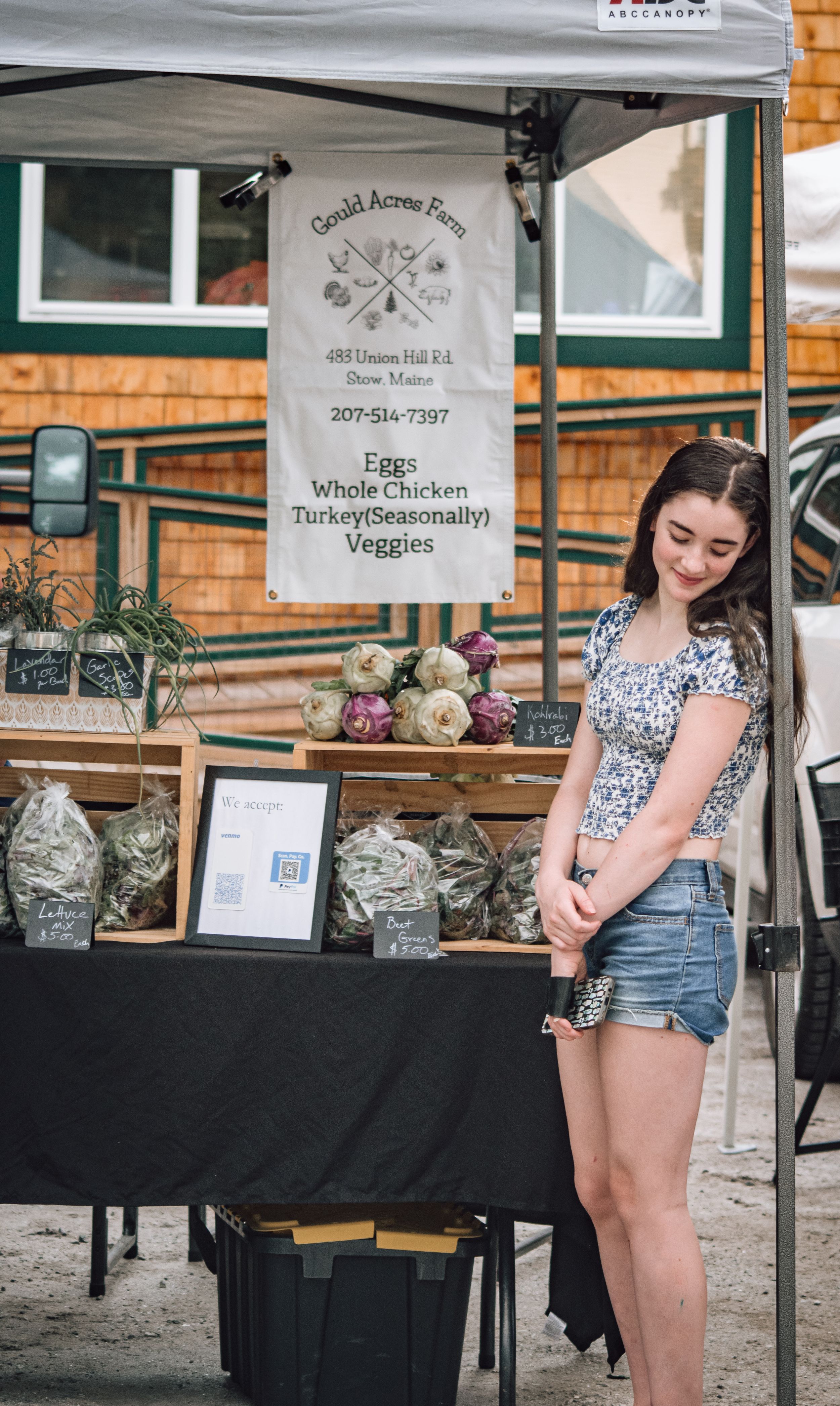 A young woman stands smiling at the Gould Acres Farm vendor booth at an outdoor market.