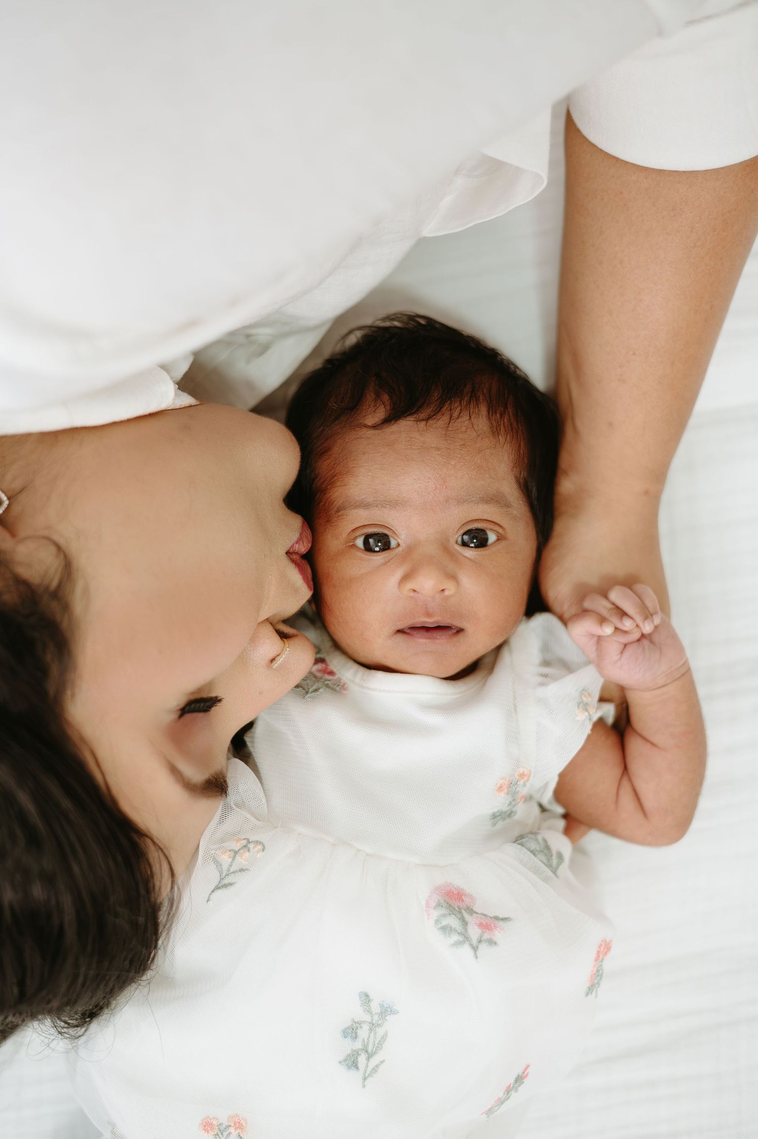 A newborn baby in a floral onesie lies on white bedding while a mother tenderly kisses their cheek.