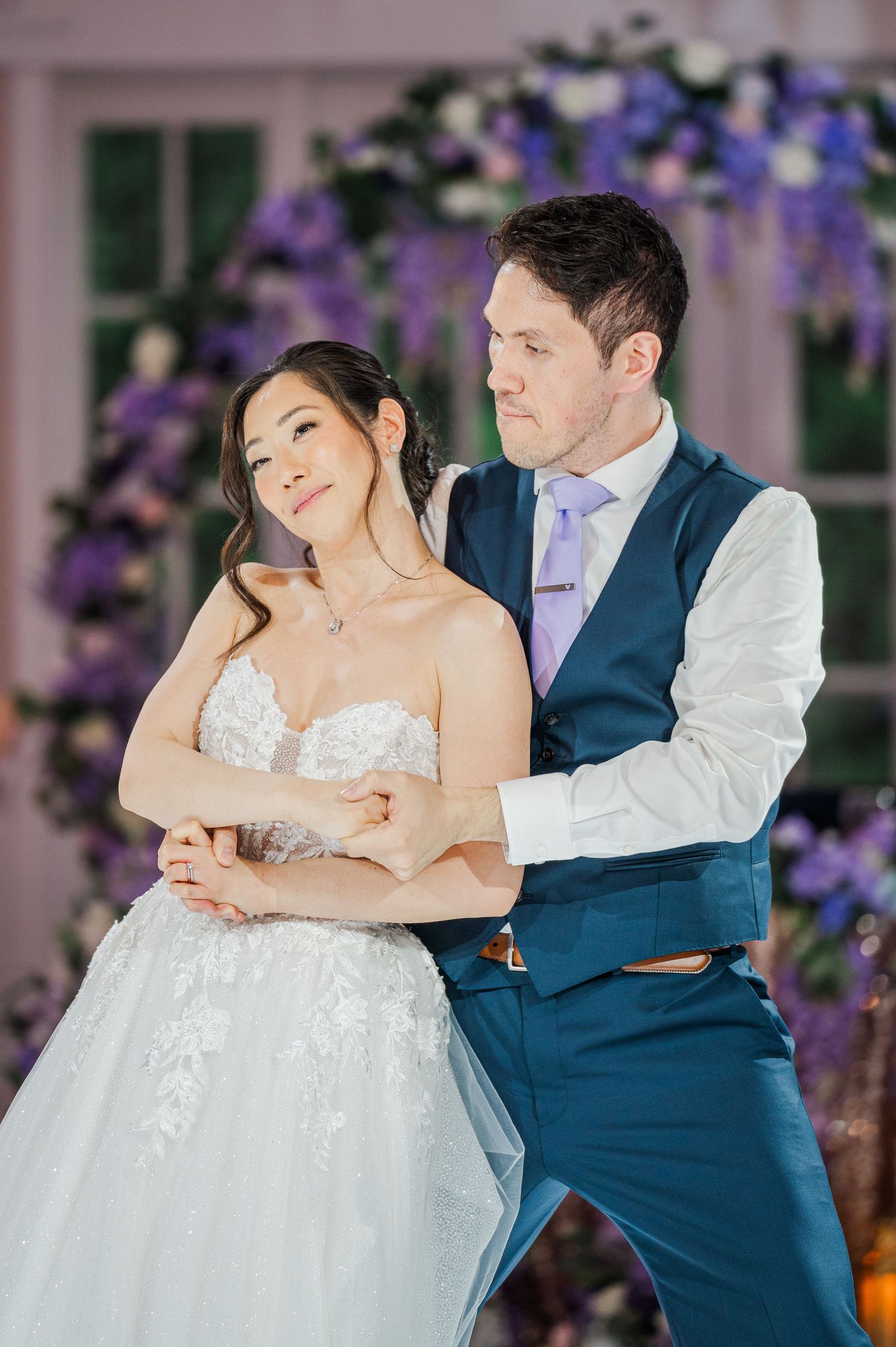Bride and groom share first dance, surrounded by purple floral arrangements at elegant wedding reception.