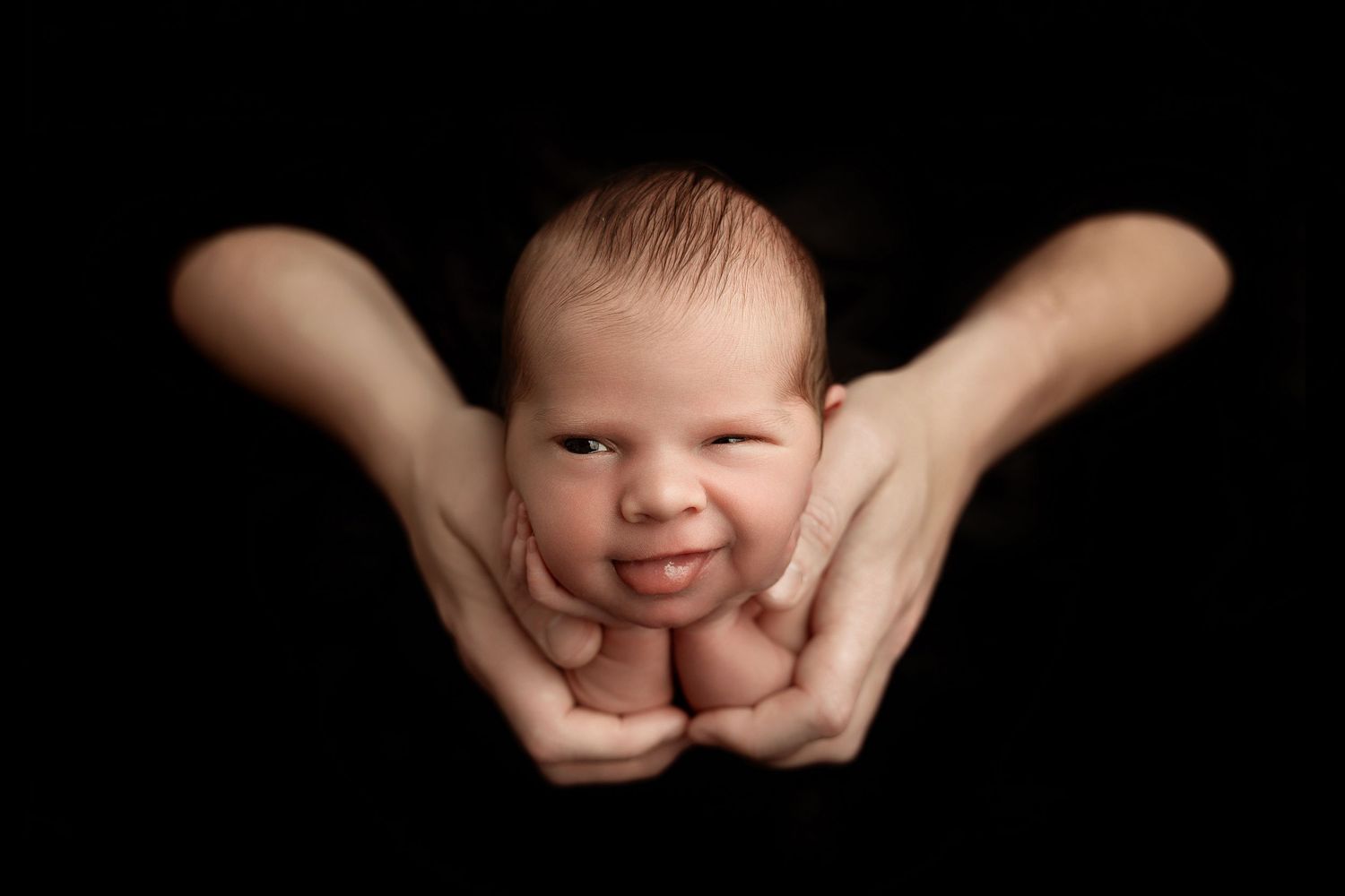 Newborn baby cradled gently in adult hands against black background, eyes open in a classic newborn portrait photo.