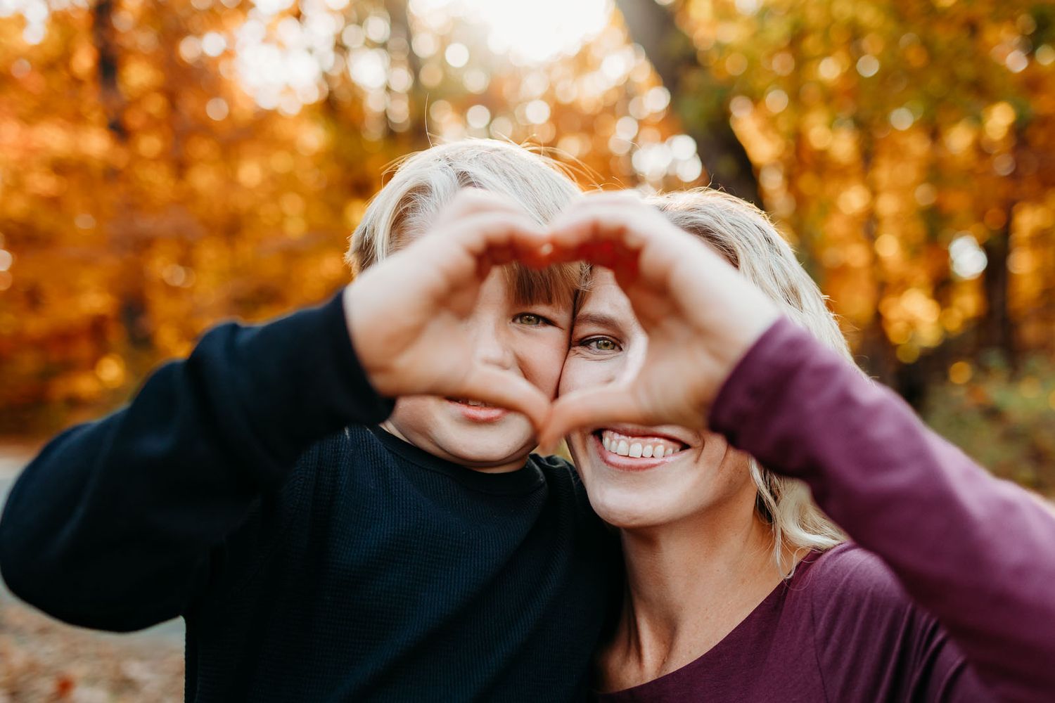 A mother and son making a heart with their hands and smiling. The trees have changed color and the sun is shining.