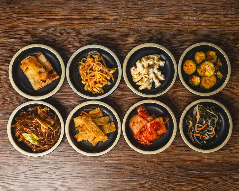 Eight small black bowls of Korean banchan side dishes arranged on a wooden table, featuring kimchi, noodles, and vegetables.