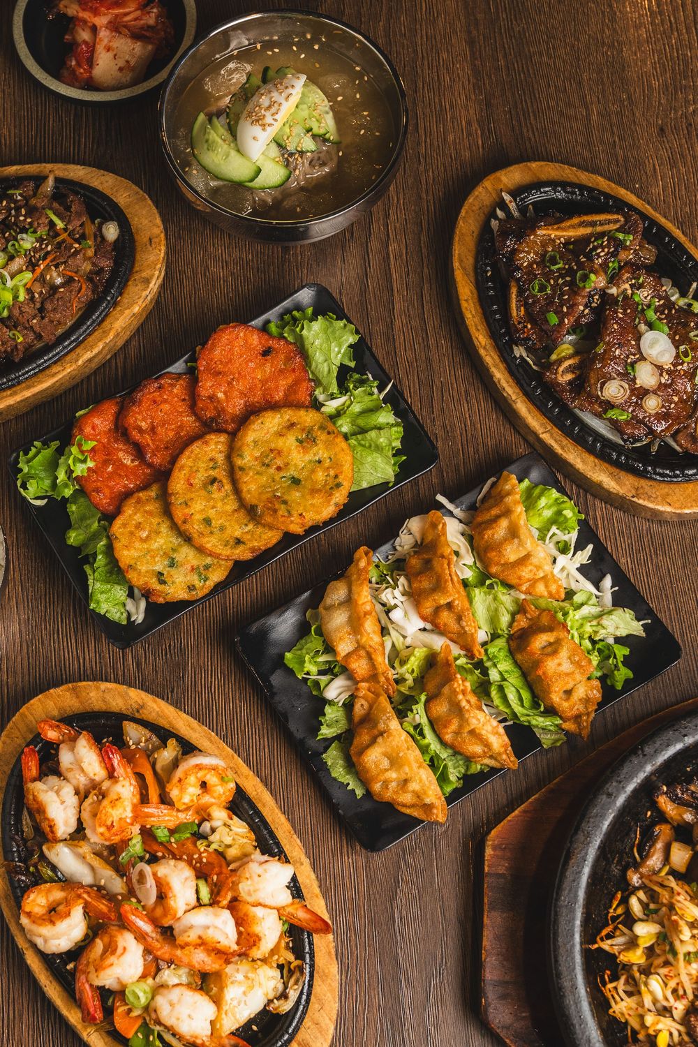 Overhead view of Korean feast with dumplings, shrimp, pancakes, octopus, and soup dishes on wooden table.
