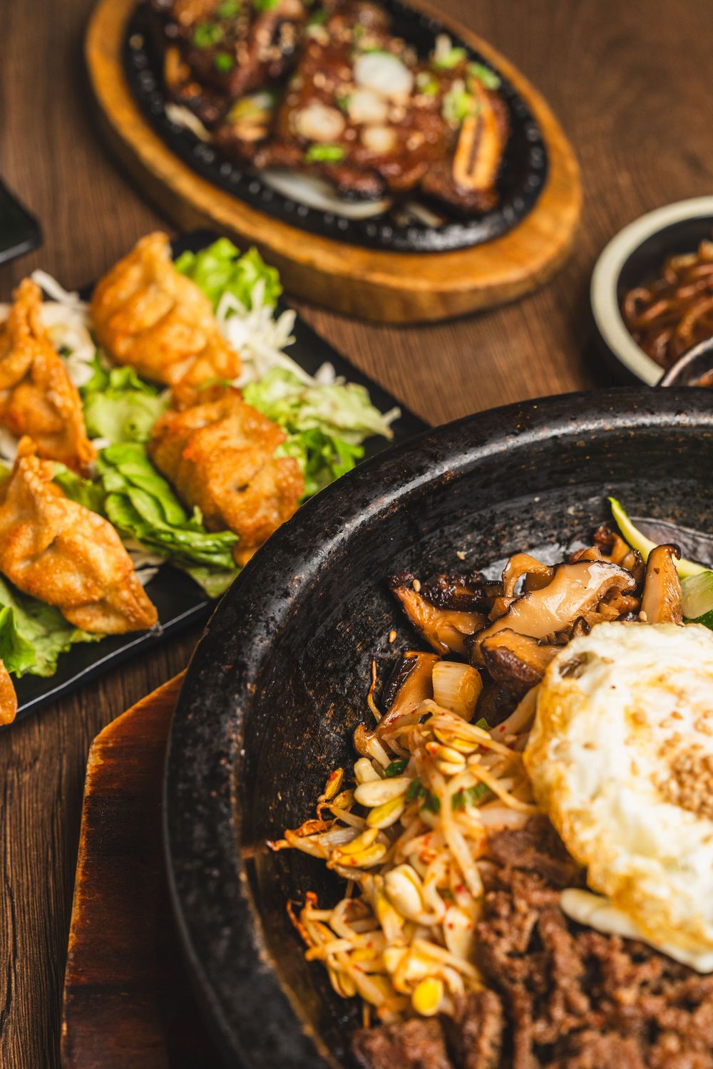 Sizzling Korean bibimbap in cast iron skillet with fried egg, mushrooms, and beef, alongside dumplings and grilled meat.