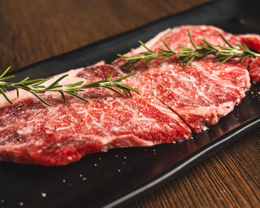 Raw beef slices seasoned with salt and topped with fresh rosemary on a black plate, ready for grilling.