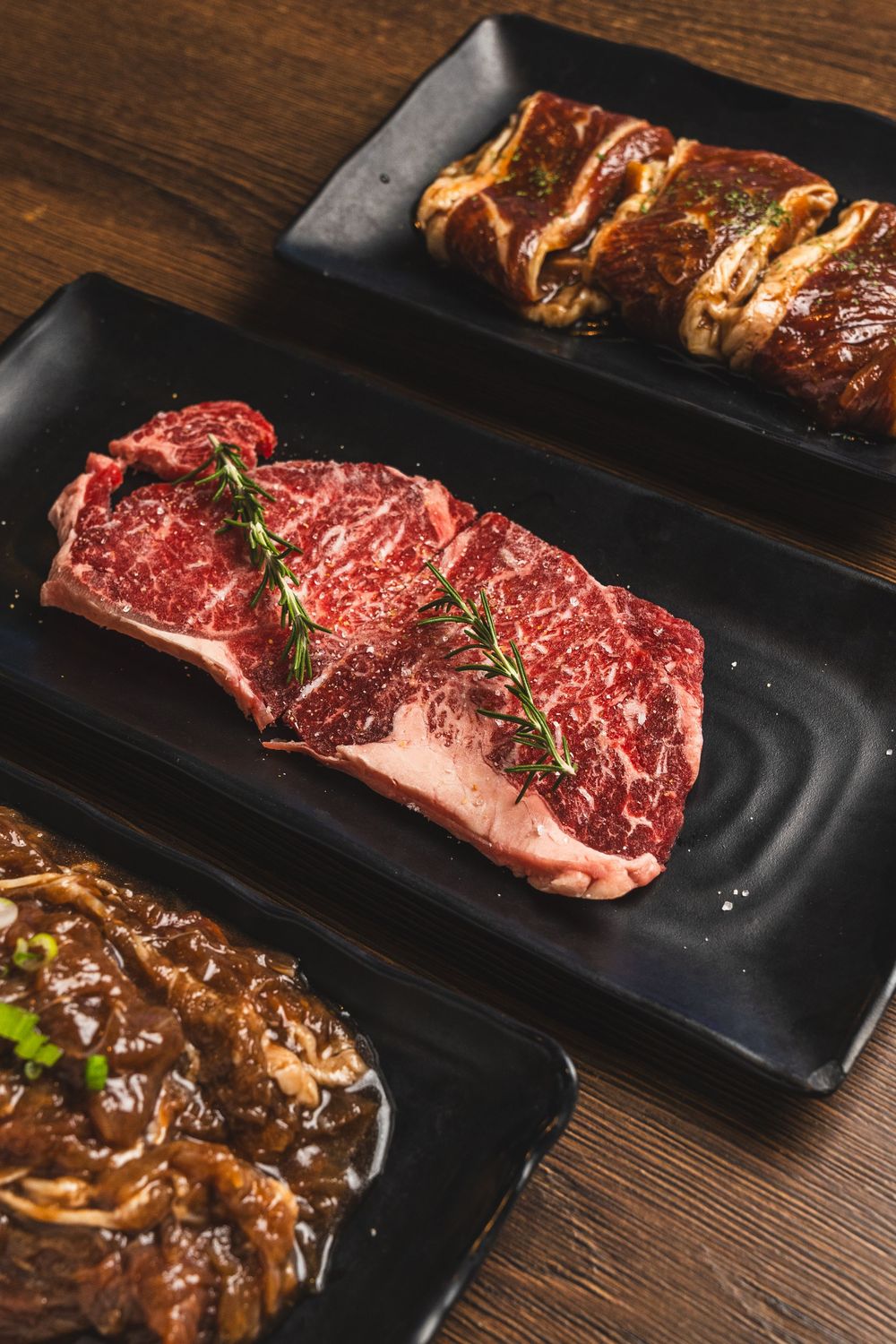 Three premium raw beef steaks with rosemary on black rectangular plates displayed on a rustic wooden table.