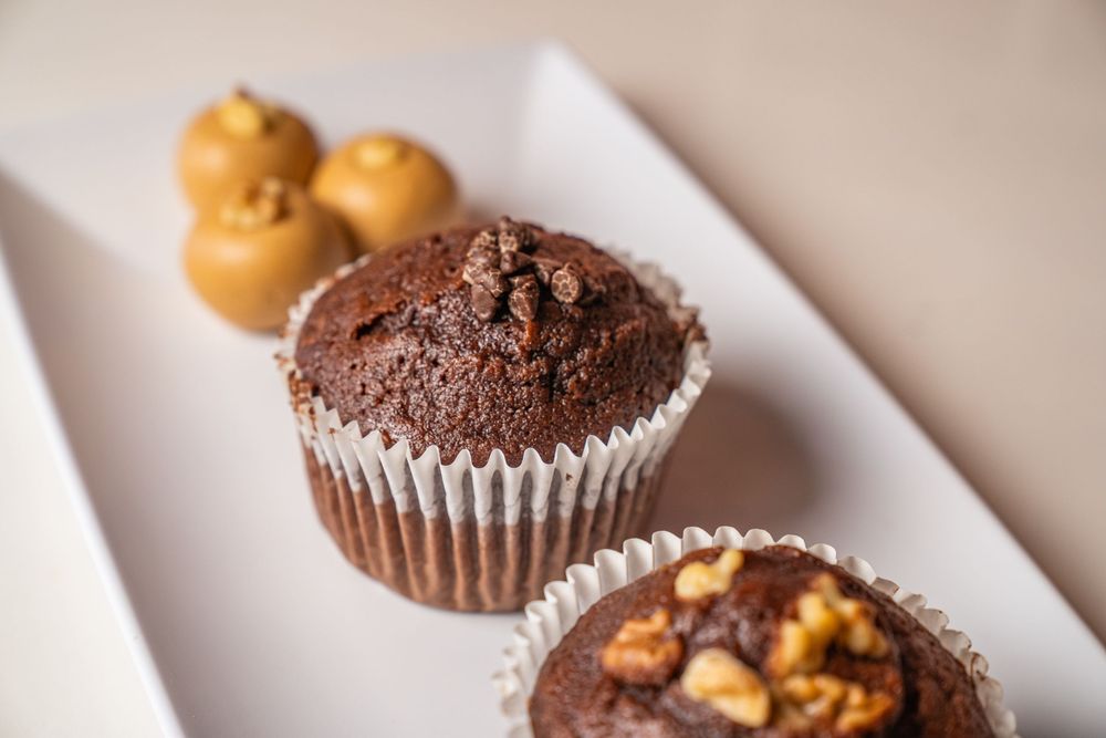 Two chocolate walnut muffins in white paper cups on a white plate, with small caramel treats in the background.
